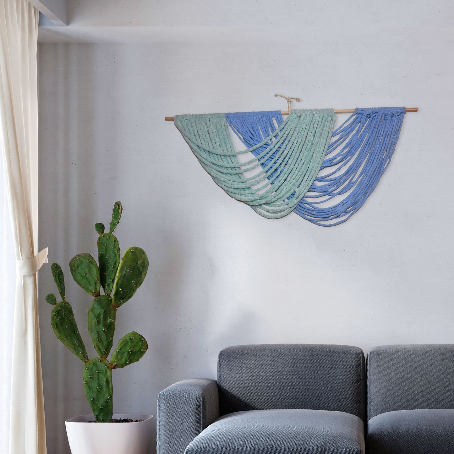 Handmade macramé wall hanging with layered arcs of green and blue yarn, draped in ocean wave-like curves from a wooden dowel.