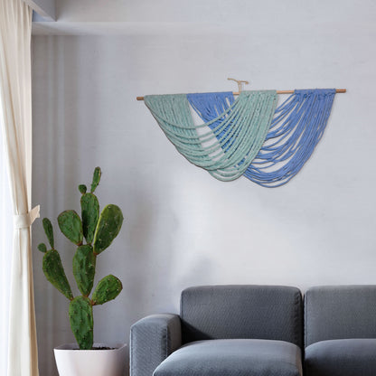 Handmade macramé wall hanging with layered arcs of green and blue yarn, draped in ocean wave-like curves from a wooden dowel.