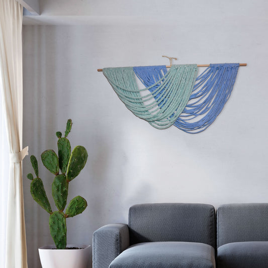 Handmade macramé wall hanging with layered arcs of green and blue yarn, draped in ocean wave-like curves from a wooden dowel.